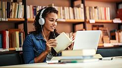 Junge Frau mit Kopfhören sitzt in einer Bibliothek vor einem Laptop und blättert in einem Buch.