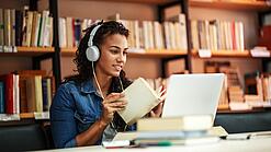 Junge Frau mit Kopfhören sitzt in einer Bibliothek vor einem Laptop und blättert in einem Buch.