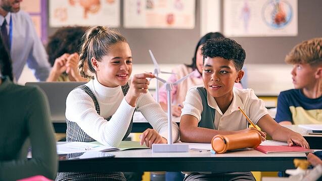 Zwei Kinder sitzen an einem Schultisch und haben ein Modell einer Windkraftturbine vor sich.