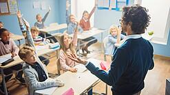 Ein junger, braunhaariger Lehrer mit einer Brille steht vor einer Klasse Schüler:innen, die sich engagiert melden.