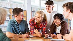 Sechs Schülerinnen und Schüler sind um einen Tisch gebeugt und diskutieren gut gelaunt über etwas.