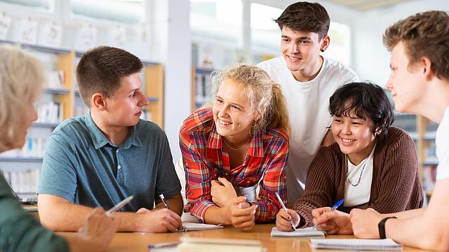 Sechs Schülerinnen und Schüler sind um einen Tisch gebeugt und diskutieren gut gelaunt über etwas.
