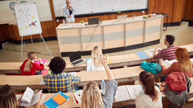 Mehrere Studierende sitzen in einem Hörsaal und schauen zur Professorin nach vorne, ein Studentin meldet sich.