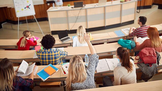 Mehrere Studierende sitzen in einem Hörsaal und schauen zur Professorin nach vorne, ein Studentin meldet sich.