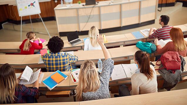 Mehrere Studierende sitzen in einem Hörsaal und schauen zur Professorin nach vorne, ein Studentin meldet sich.