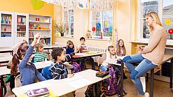 Junge Lehrerin sitzt vor einer Grundschulklasse in der mehrere Kinder aufzeigen.
