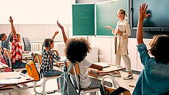 Grundschulkinder sitzen in einem Klassenzimmer und heben die Hände zur Wortmeldung, an der Tafel steht eine Lehrerin.