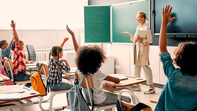 Grundschulkinder sitzen in einem Klassenzimmer und heben die Hände zur Wortmeldung, an der Tafel steht eine Lehrerin.
