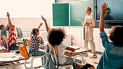 Grundschulkinder sitzen in einem Klassenzimmer und heben die Hände zur Wortmeldung, an der Tafel steht eine Lehrerin.