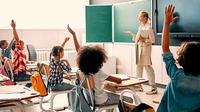Grundschulkinder sitzen in einem Klassenzimmer und heben die Hände zur Wortmeldung, an der Tafel steht eine Lehrerin.