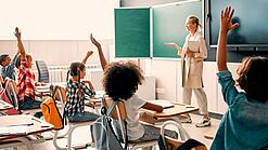 Grundschulkinder sitzen in einem Klassenzimmer und heben die Hände zur Wortmeldung, an der Tafel steht eine Lehrerin.