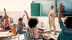 Grundschulkinder sitzen in einem Klassenzimmer und heben die Hände zur Wortmeldung, an der Tafel steht eine Lehrerin.
