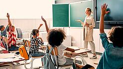 Grundschulkinder sitzen in einem Klassenzimmer und heben die Hände zur Wortmeldung, an der Tafel steht eine Lehrerin.