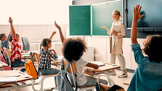 Grundschulkinder sitzen in einem Klassenzimmer und heben die Hände zur Wortmeldung, an der Tafel steht eine Lehrerin.