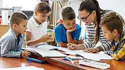 Eine Lehrerin ist umringt von Schülern und korrigiert Schulaufgaben.