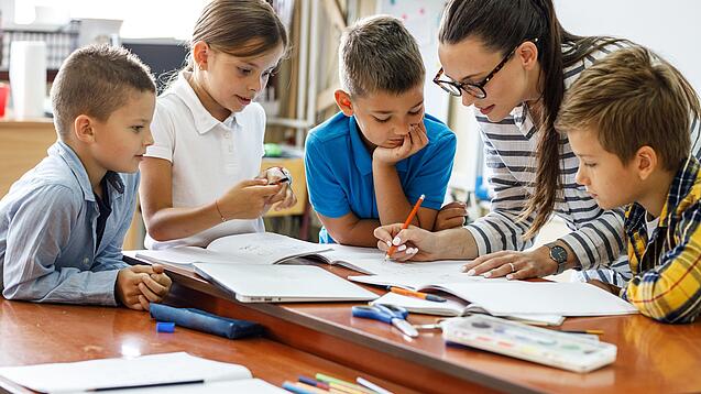 Eine Lehrerin ist umringt von Schülern und korrigiert Schulaufgaben.