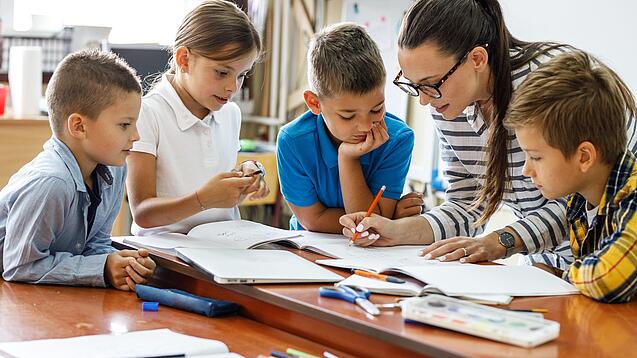 Eine Lehrerin ist umringt von Schülern und korrigiert Schulaufgaben.