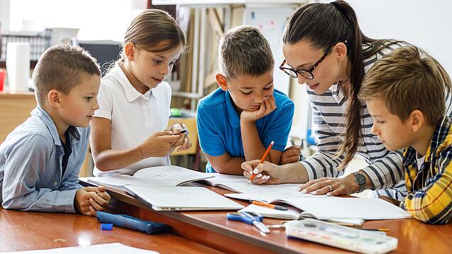 Eine Lehrerin ist umringt von Schülern und korrigiert Schulaufgaben.
