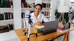 Frau sitzt am Schreibtisch vor einem Laptop und einem Handystativ.