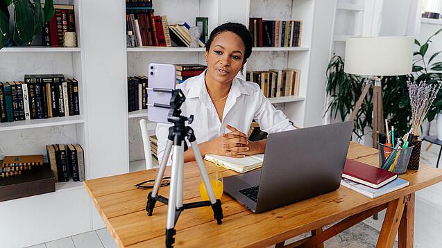 Frau sitzt am Schreibtisch vor einem Laptop und einem Handystativ.