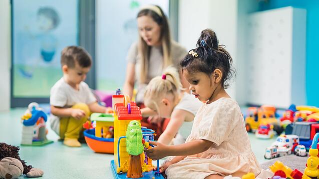 Kinder sitzen mit einer Erzieherin auf dem Boden und spielen.