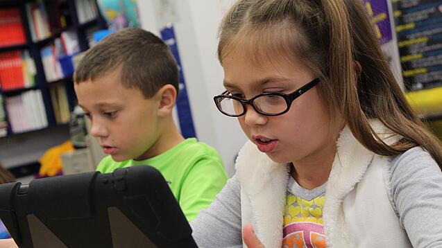 Zwei Kinder sitzen an einem Tisch und schauen auf ein Tablet.