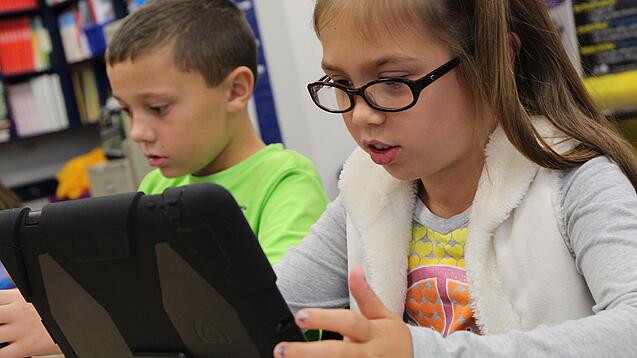 Zwei Kinder sitzen an einem Tisch und schauen auf ein Tablet.