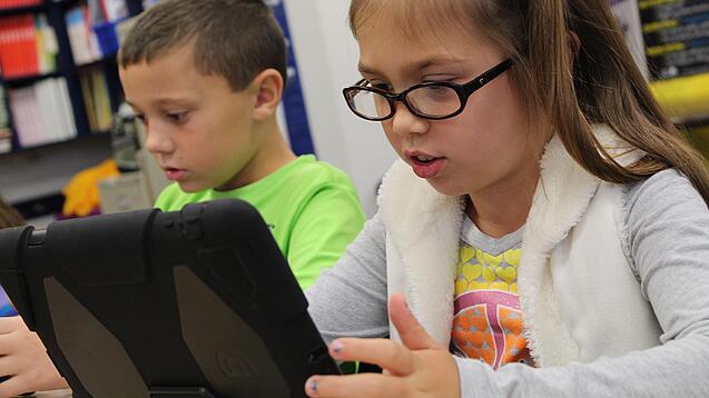 Zwei Kinder sitzen an einem Tisch und schauen auf ein Tablet.