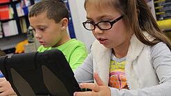 Zwei Kinder sitzen an einem Tisch und schauen auf ein Tablet.