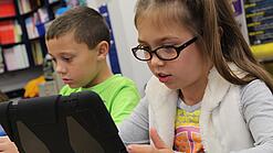 Zwei Kinder sitzen an einem Tisch und schauen auf ein Tablet.