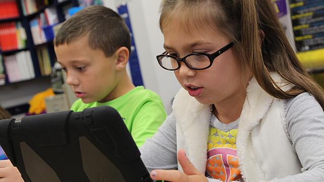 Zwei Kinder sitzen an einem Tisch und schauen auf ein Tablet.