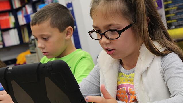 Zwei Kinder sitzen an einem Tisch und schauen auf ein Tablet.