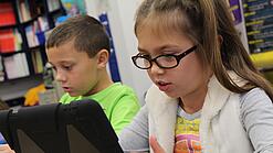 Zwei Kinder sitzen an einem Tisch und schauen auf ein Tablet.
