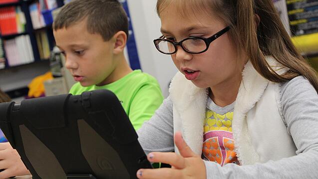 Zwei Kinder sitzen an einem Tisch und schauen auf ein Tablet.
