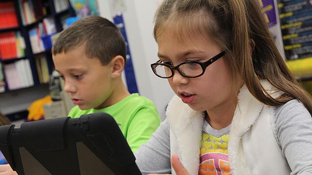 Zwei Kinder sitzen an einem Tisch und schauen auf ein Tablet.
