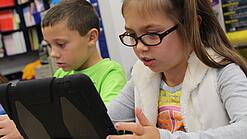 Zwei Kinder sitzen an einem Tisch und schauen auf ein Tablet.