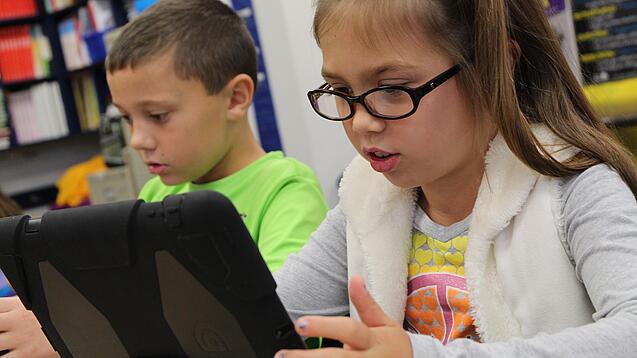 Zwei Kinder sitzen an einem Tisch und schauen auf ein Tablet.