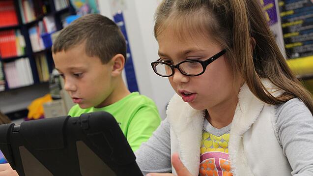 Zwei Kinder sitzen an einem Tisch und schauen auf ein Tablet.