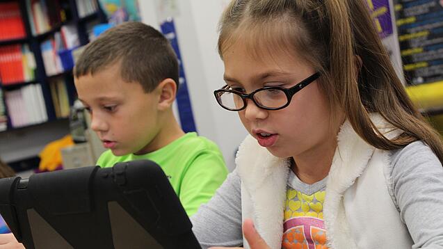 Zwei Kinder sitzen an einem Tisch und schauen auf ein Tablet.