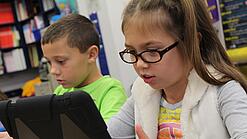 Zwei Kinder sitzen an einem Tisch und schauen auf ein Tablet.