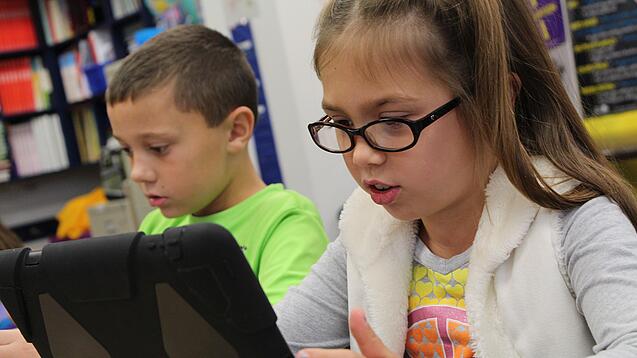 Zwei Kinder sitzen an einem Tisch und schauen auf ein Tablet.