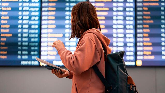 Junge Frau steht vor einer Anzeigetafel an einem Flughafen.