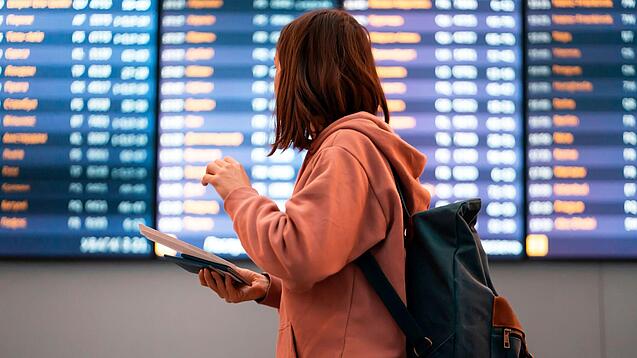 Junge Frau steht vor einer Anzeigetafel an einem Flughafen.