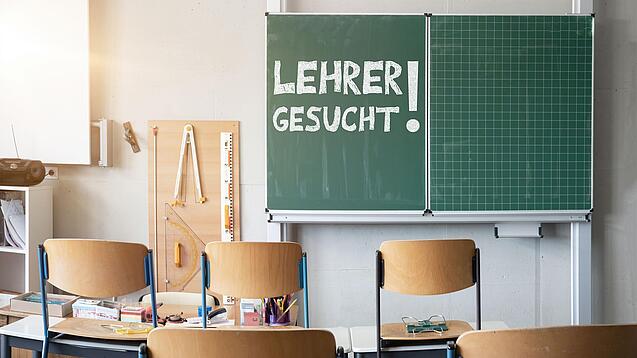 Auf einer Tafel in einem Klassenzimmer steht groß mit Kreide "Lehrer gesucht!" geschrieben.