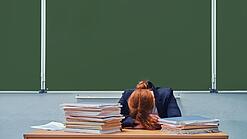 Frau sitzt an einem Lehrerpult vor einer Tafel und hat den Kopf auf dem Pult abgelegt.