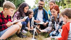 Eine Gruppe Schüler:innen sitzt mit zwei Lehrkräften um das Modell eines Windrads.