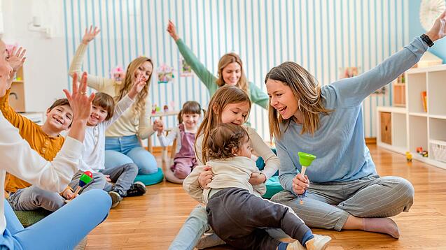 Erzieherinnen sitzen mit Kindergartenkindern auf dem Boden und singen.