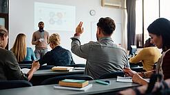 Mehrere Menschen sitzen in einem Seminarraum und schauen auf eine Präsentation, eine Person meldet sich.