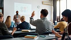 Mehrere Menschen sitzen in einem Seminarraum und schauen auf eine Präsentation, eine Person meldet sich.