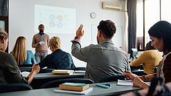 Mehrere Menschen sitzen in einem Seminarraum und schauen auf eine Präsentation, eine Person meldet sich.